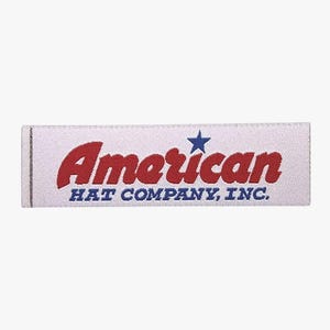 May include: A white rectangular label featuring the word "American" in red, a blue star, and the text "HAT COMPANY, INC." in blue. The label has a woven texture and a grey border on the left.