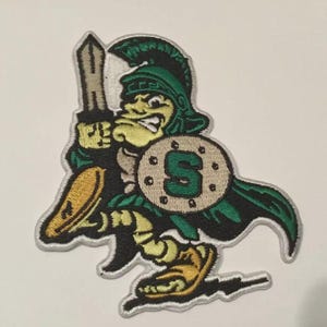 Op de afbeelding: Geborduurde patch met een cartoon Spartaanse krijger in groen, geel en zwart. De krijger houdt een zwaard en een schild met de letter "S" vast. De patch heeft een witte rand.