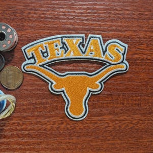May include: Embroidered patch with the word "TEXAS" in gold lettering and a black outline, above a gold longhorn silhouette. The patch has a white border. A penny and sewing supplies are in the background, set against a wooden surface.