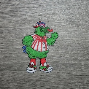 May include: Embroidered patch of a green cartoon creature with a red tongue, wearing a red and white striped shirt, red socks, and trainers. The creature has a colourful hat and a friendly pose, ideal for adding to clothing or accessories.