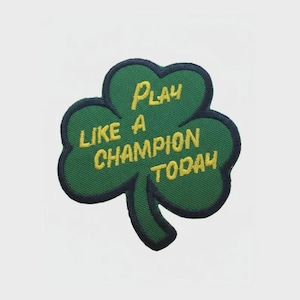 May include: A green shamrock-shaped patch with the words "PLAY LIKE A CHAMPION TODAY" embroidered in yellow. It has a dark blue border and a white background.