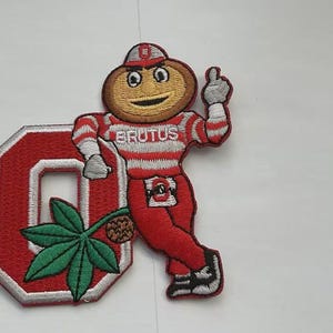 May include: Embroidered patch featuring the mascot Brutus Buckeye in a red and white striped outfit, giving a thumbs-up. The patch includes a red "O" and a green leaf with a brown nut. The word "BRUTUS" is across the chest.