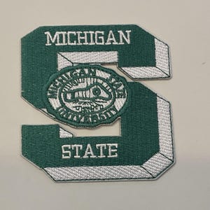 Op de afbeelding: Een groen en wit geborduurde patch in de vorm van de letter "S". De patch heeft de woorden "MICHIGAN" en "STATE" in witte letters. Een embleem met de woorden "MICHIGAN STATE UNIVERSITY" staat in het midden.