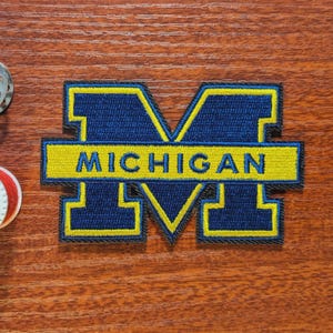 Peut inclure: Écusson brodé avec la lettre "M" bleu marine, contour jaune et le mot "MICHIGAN" en jaune. Le patch est rectangulaire avec une bordure noire. Idéal pour vêtements ou accessoires.
