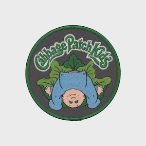 May include: Embroidered patch featuring a Cabbage Patch Kids design. The patch has a circular shape with a dark gray background and a green border. The design includes a baby in a blue outfit, green leaves, and the words "Cabbage Patch Kids" in green.