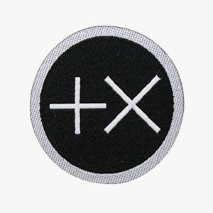 May include: A circular patch with a black center and a white border. The black center features a white plus sign and a white X, creating a minimalist design. The patch is likely for clothing or accessories.