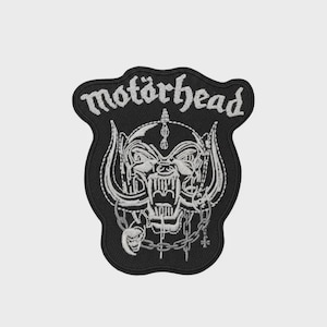 Puede incluir: Parche bordado negro con la palabra "motörhead" en escritura blanca sobre una ilustración detallada de una calavera con colmillos, cuernos y cadenas. El parche tiene un borde negro, ideal para ropa o accesorios.