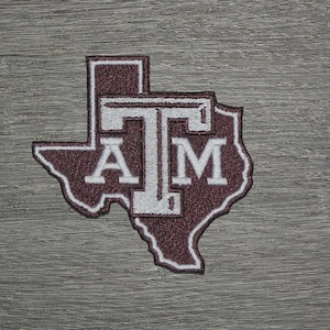 May include: Maroon and white embroidered patch in the shape of Texas, featuring the letters "A" and "M" stacked below a "T". The patch has a white border and is set against a gray, textured background.