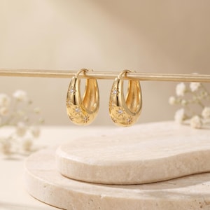 May include: A pair of gold-colored hoop earrings. Each earring features a series of star-shaped designs with small, clear gemstones. The earrings are displayed on a gold-colored rod against a neutral background.