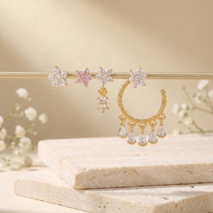 May include: A selection of gold-coloured earrings on a gold bar. The earrings showcase floral and teardrop designs, adorned with clear and pink gemstones. The earrings are presented against a neutral backdrop with a stone-like surface.