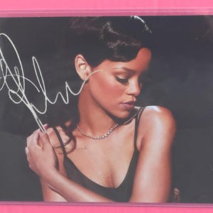 May include: A photograph of a celebrity with a signature. The image shows a woman with dark hair and a black dress, wearing a necklace. The background is dark. The signature is in white.