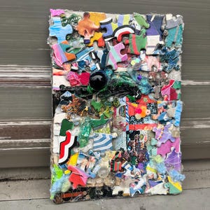 Puede incluir: Una obra de arte rectangular de técnica mixta compuesta por piezas de rompecabezas coloridas. Las piezas están dispuestas en un patrón de mosaico, con un objeto esférico negro y varias texturas. Los bordes son irregulares, lo que le da un aspecto único y hecho a mano.