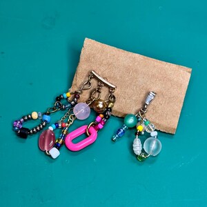 Puede incluir: Dos pendientes hechos a mano con cuentas y abalorios de colores. Un pendiente presenta un dije ovalado rosa, mientras que el otro tiene un grupo de cuentas verdes y transparentes. Los pendientes están sobre una tarjeta marrón sobre un fondo verde.