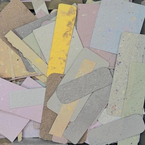 May include: A pile of assorted paper shapes in pastel shades of yellow, pink, grey and beige. Some pieces have speckled patterns, and the edges are torn or uneven, suggesting a handmade or recycled origin. Metric measurements.