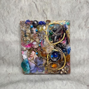 May include: A mixed media art piece with a square shape. The artwork features a collage of various materials, including beads, gems, and metallic elements. The color palette includes gold, blue, pink, and clear accents, creating a textured and layered effect.