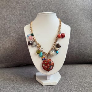 Puede incluir: Un collar de cadena dorada con varios abalorios, incluyendo cuentas y un colgante redondo rojo con un diseño floral. El collar se exhibe en un busto de joyería blanco sobre un fondo gris.