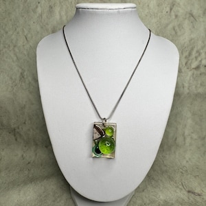 Puede incluir: Collar con colgante rectangular y cadena plateada. El colgante transparente contiene elementos verdes y blancos, incluyendo esferas. El collar se exhibe en un maniquí blanco.
