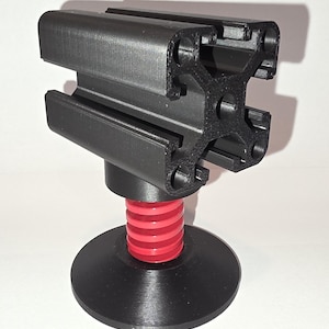 May include: A black, 3D-printed aluminum extrusion profile mounted on a red and black base. The profile has multiple slots and holes. The base features a red, ribbed section and a black, circular foot. The object is designed for structural support.