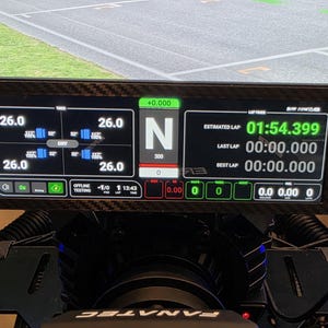 May include: A digital racing display with a carbon fiber frame. The screen shows lap times, weather conditions, and other telemetry data. The estimated lap time is 01:54.399. The display is mounted on a racing simulator.