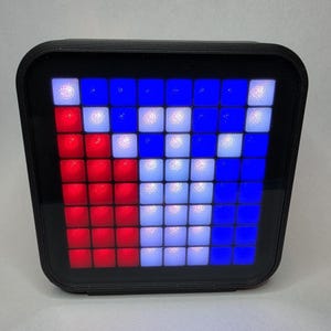 Puede incluir: Una pantalla digital negra de forma cuadrada con una cuadrícula de cuadrados iluminados en rojo, blanco y azul. La pantalla recuerda a una bandera o a un diseño de arte pixelado retro. El dispositivo tiene un borde negro.