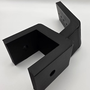 Puede incluir: Un soporte negro en forma de L con una abertura rectangular y varios agujeros pretaladrados. El soporte está hecho de un material mate sólido y parece estar diseñado para soporte estructural o montaje. El diseño incluye un agujero circular en el lateral.