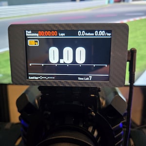 Może przedstawiać: Czarny cyfrowy wyświetlacz pokazujący dane z wyścigu, w tym pozostałe paliwo, czas okrążenia i galony. Ekran wyraźnie wyświetla "0.00". Urządzenie jest zamontowane na czarnej podstawie z widocznym napisem "NATEC".