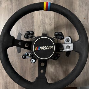 May include: A black racing steering wheel with a circular center hub featuring the NASCAR logo in white, yellow, red, and blue. The wheel has a black suede-like grip with a yellow, red, and blue stripe at the top, and includes integrated buttons.