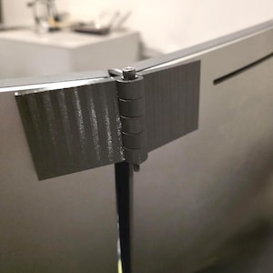 Puede incluir: Una bisagra gris oscuro, impresa en 3D, fijada a una superficie curva de color gris claro. La bisagra está compuesta por múltiples segmentos cilíndricos conectados por un tornillo. La superficie tiene un aspecto texturizado.