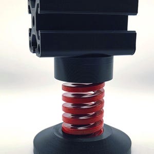 Op de afbeelding: Een zwart, industrieel veerelement. Het heeft een zwarte basis, een rode spiraalveer en een zwart bovendeel met een rechthoekig profiel. Het ontwerp suggereert een mechanische of technische toepassing.
