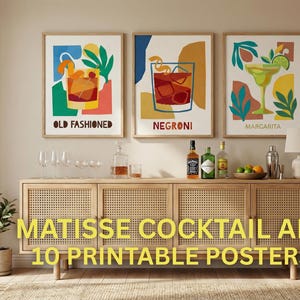 Op de afbeelding: Drie ingelijste kunstprints met cocktailillustraties: een Old Fashioned, een Negroni en een Margarita. De prints staan boven een houten kast met flessen, glazen en een plant. De tekst "MATISSE COCKTAIL ART 10 PRINTABLE POSTERS" staat onderaan.
