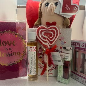 May include: A gift set featuring a plush animal, a heart-shaped lollipop, a bottle of perfume, and a lip jelly. The set includes a card that reads "Love is a Blessing" and a box of fragrance duo. The items are in shades of red, pink, and white.