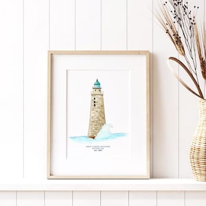 May include: A framed watercolor print of Minot's Ledge Lighthouse in Scituate, MA. The lighthouse is depicted in beige and gray tones, with a teal top, set against a blue ocean and white wave. The print is in a light wood frame.