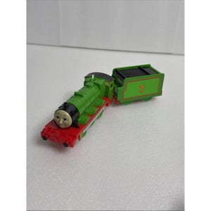 May include: A green toy steam locomotive with a red chassis and a black front face featuring a smiling expression. Attached is a green coal tender marked with the number 3 in orange. The train engine has a detailed boiler, cab, and wheels.