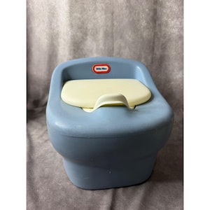 May include: A blue plastic potty training chair with a cream-colored lid and a small molded splash guard. The front of the chair features a red, white, and blue Little Tikes brand logo centered above the seat. The design is smooth and rounded.