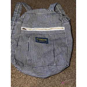 May include: A navy blue and white pinstriped backpack featuring a horizontal zippered front pocket and a rectangular navy blue patch with the yellow text OshKosh B'gosh The Genuine Article centered on the front of the bag with two shoulder straps.