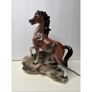 May include: A glossy ceramic horse figurine lamp featuring a brown body with a black mane and tail. The horse stands on a raised, textured base with muted green and grey tones. A thin electrical cord extends from the back of the base.