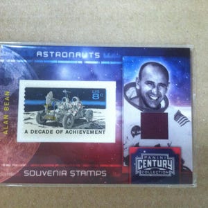 May include: A souvenir stamp card featuring a U.S. 8¢ stamp with a lunar rover and the text "A Decade of Achievement." The card also includes a portrait of astronaut Alan Bean and the text "Astronauts" and "Souvenir Stamps."