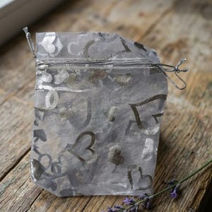 Op de afbeelding: Een semi-transparant, lichtgrijs organza zakje met trekkoord, voorzien van een herhalend patroon van metallic zilveren hartjes. Het zakje is gemaakt van een lichtgewicht, doorschijnende stof en heeft dunne zilveren koordjes als sluiting.