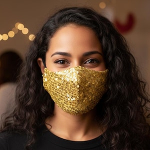Puede incluir: Primer plano de una persona con una mascarilla dorada con lentejuelas. La mascarilla cubre la nariz y la boca. La persona tiene el pelo rizado oscuro y está sonriendo. El fondo está borroso, con luces de cadena y otras personas visibles.