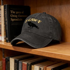 May include: Dark gray baseball cap with a curved brim. The cap features the text "F-CAW-F" in gold lettering above a black raven illustration. The cap is sitting on a wooden shelf with books.