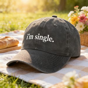 May include: A dark gray baseball cap with the text "i'm single." in white. The cap is sitting on a picnic blanket with a loaf of bread and a basket of flowers in the background. The cap has a curved brim and a button on top.