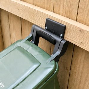 Op de afbeelding: Een groene plastic afvalbak met een zwarte montagebeugel bevestigd aan een houten schutting. De bak is voorzien van een scharnierend deksel en een handvat, met de tekst WASTE ONLY en een pijl in het wit op het deksel gedrukt.