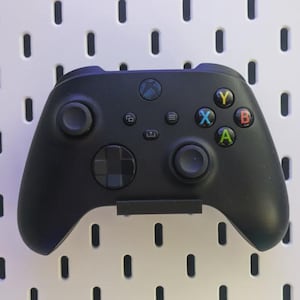 May include: A black Xbox controller with colored buttons and a directional pad. The controller has two analog sticks, a D-pad, and several function buttons. The controller is set against a white background with evenly spaced holes.