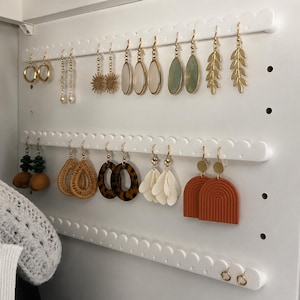 Puede incluir: Un organizador de pendientes de pared en color blanco con tres barras horizontales festoneadas y orificios perforados para colgar diversos estilos de pendientes, incluyendo aros dorados, colgantes de hojas, arcos texturizados y cuentas.