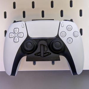 Puede incluir: Un mando inalámbrico PlayStation 5 DualSense blanco y negro, con un soporte negro. El mando tiene un diseño simétrico con un panel táctil, botones y joysticks. El fondo es un panel perforado blanco.