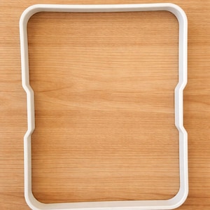 May include: A white, square cookie cutter with rounded corners and a notched design on two sides. The cutter is made of plastic and sits on a light brown wooden surface.
