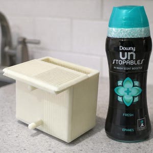 Peut inclure: Une bouteille noire de booster de parfum de lavage Downy Unstopables avec un bouchon turquoise et un motif floral turquoise. À côté, un distributeur de booster de parfum de lessive rectangulaire de couleur crème avec un couvercle.
