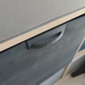 May include: Close-up of a dark grey fabric storage bin with a matching handle. The bin is part of a larger storage unit with a light wood trim and a grey top. The handle is a curved, rectangular shape.