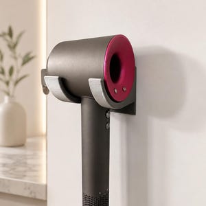 May include: A dark gray hair dryer with a vibrant magenta circular head is securely held in a light gray, U-shaped wall-mounted holder. The hair dryer features a long cylindrical handle with a mesh base and two small circular buttons on the front.
