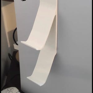 Puede incluir: Un soporte blanco para mando de consola. El soporte está diseñado para sujetar el mando y la consola. El soporte está fijado al lateral de la consola. La consola es blanca y tiene una base negra.